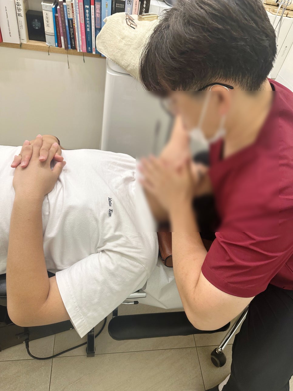 한 사람이 마사지 테이블에 누워 있고, 다른 사람이 마사지를 하고 있는 모습. 주변에 책이 진열되어 있다.