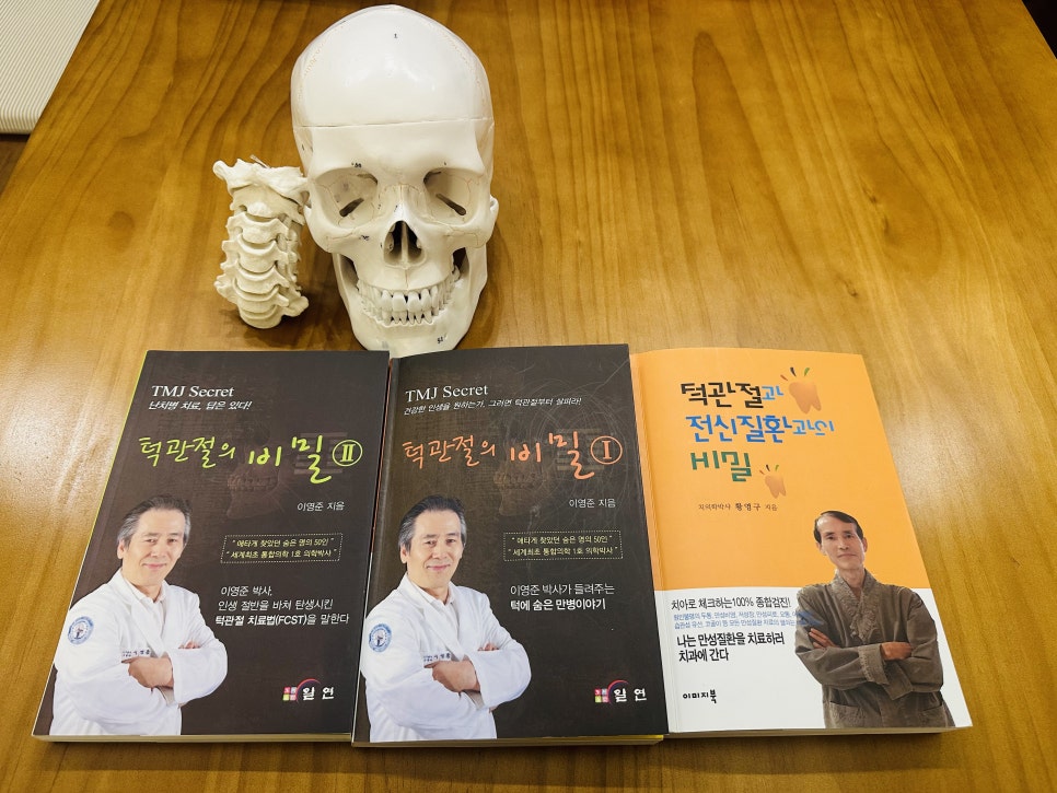 책상 위에 두개골 모형과 턱관절 관련 서적 세 권이 놓여 있다. 책 표지에는 저자의 사진과 함께 '턱관절의 비밀'이라는 제목이 적혀 있다.