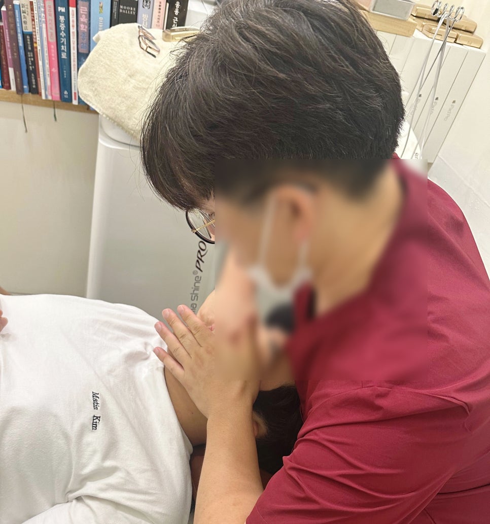 한 사람이 누워 있고, 다른 사람이 얼굴을 손으로 만지고 있는 장면입니다. 주변에 책과 의료 도구가 보입니다.
